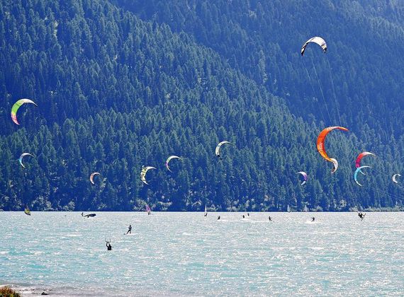 Mehrere Kitesurfer fahren auf dem Reschensee vor einem bewaldeten Berghang.