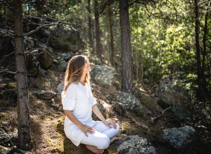Eine Frau sitzt in weißer Kleidung auf Felsen im Wald und meditiert im Sonnenlicht.