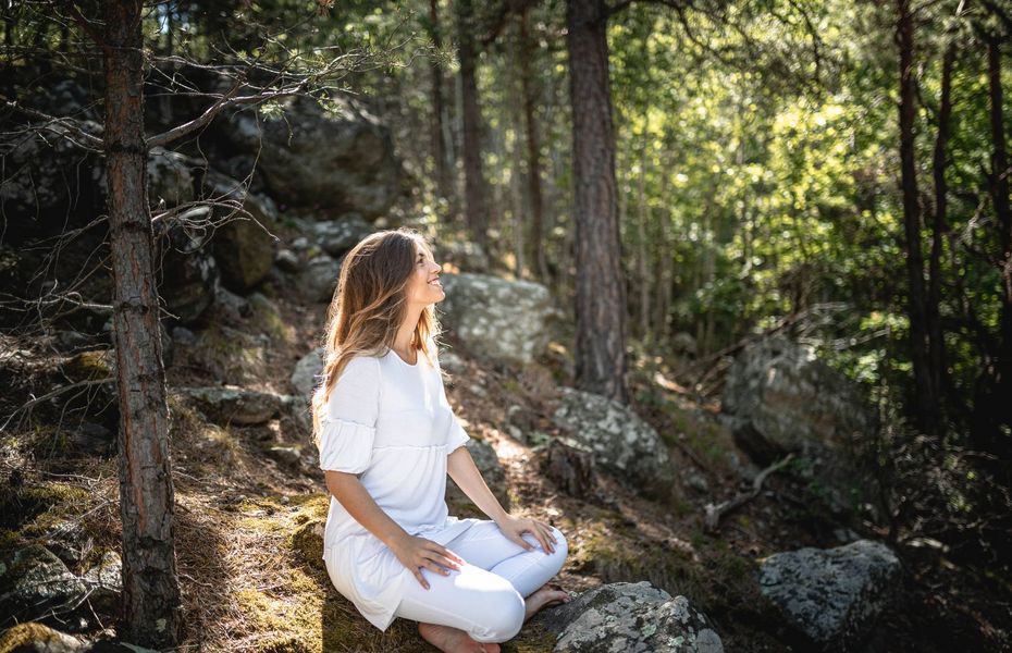 Meditation im Wald beim Preidlhof in Naturns Eine Frau sitzt in weißer Kleidung auf Felsen im Wald und meditiert im Sonnenlicht.