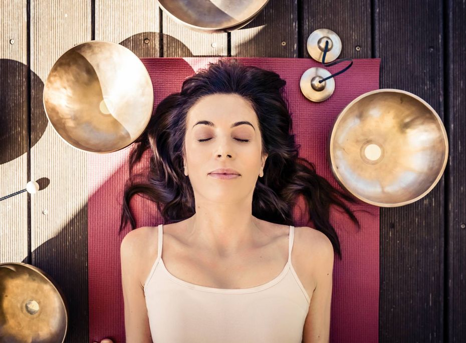 Meditation im Wellnesshotel Preidlhof in Naturns Eine Frau liegt mit geschlossenen Augen auf einer Yogamatte, umgeben von Klangschalen.