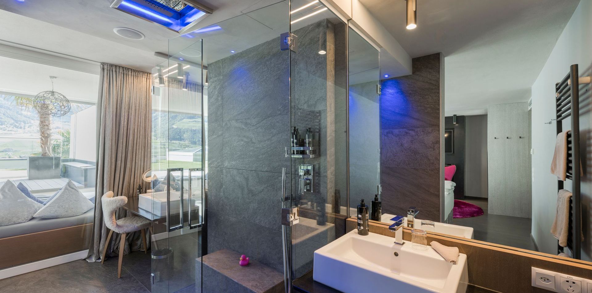 Modernes Bad einer Private Suite mit Glasdusche, Waschtisch und Blick in den Schlafbereich mit Sitznische.