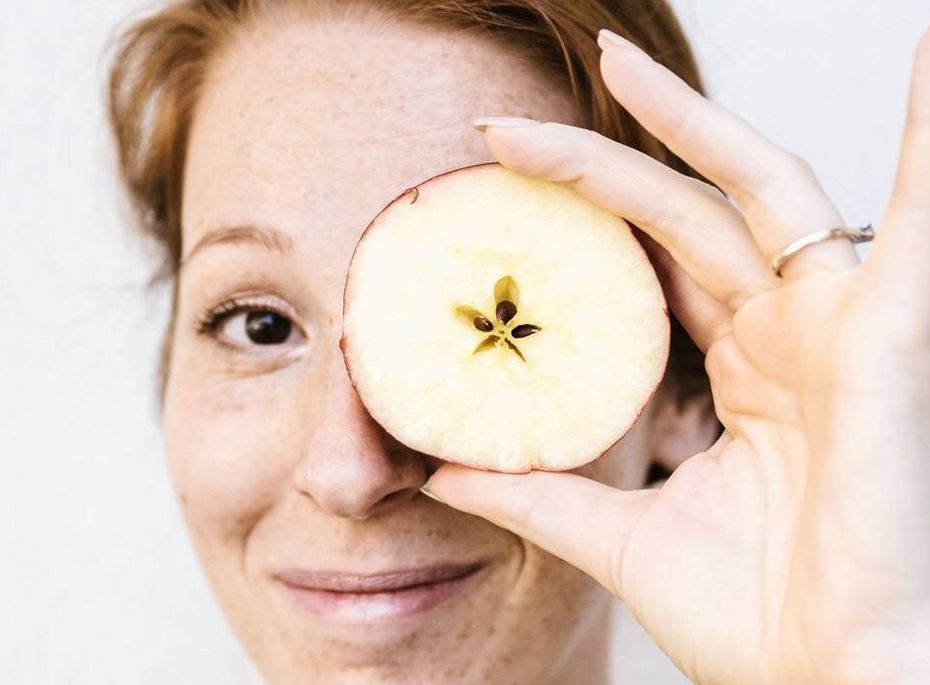 Wellnessbehandlung mit Apfel im Preidlhof Eine Frau hält eine Apfelscheibe vor ein Auge und blickt lächelnd in die Kamera.