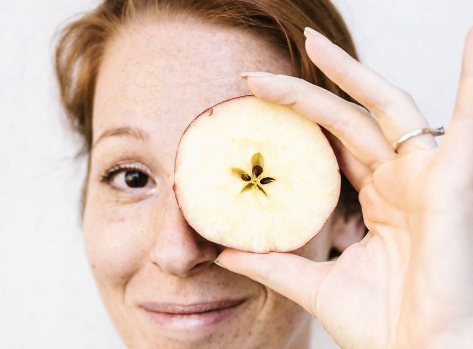 Wellnessbehandlung mit Apfel im Preidlhof Eine Frau hält eine Apfelscheibe vor ein Auge und blickt lächelnd in die Kamera.
