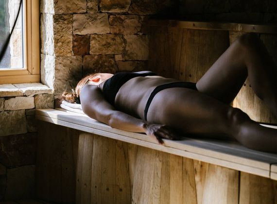 A person lies on a wooden bench in a sauna room beside a small window and stone wall.