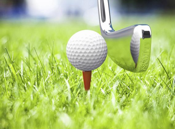 Ein Golfball liegt auf einem Tee im kurzen Gras, daneben berührt ein Golfeisen den Ball.
