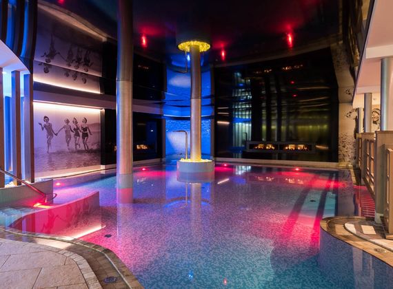 An indoor wellness pool with colored lighting, a central waterfall feature and lounge area at Preidlhof.