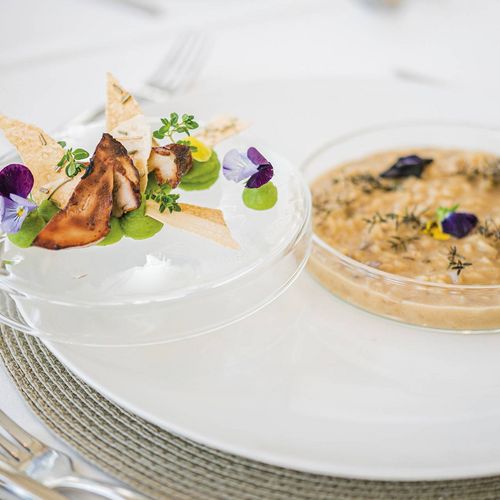 Ein fein angerichtetes Dessert und eine cremige Suppe auf weißem Geschirr mit essbaren Blüten und Kräutern.