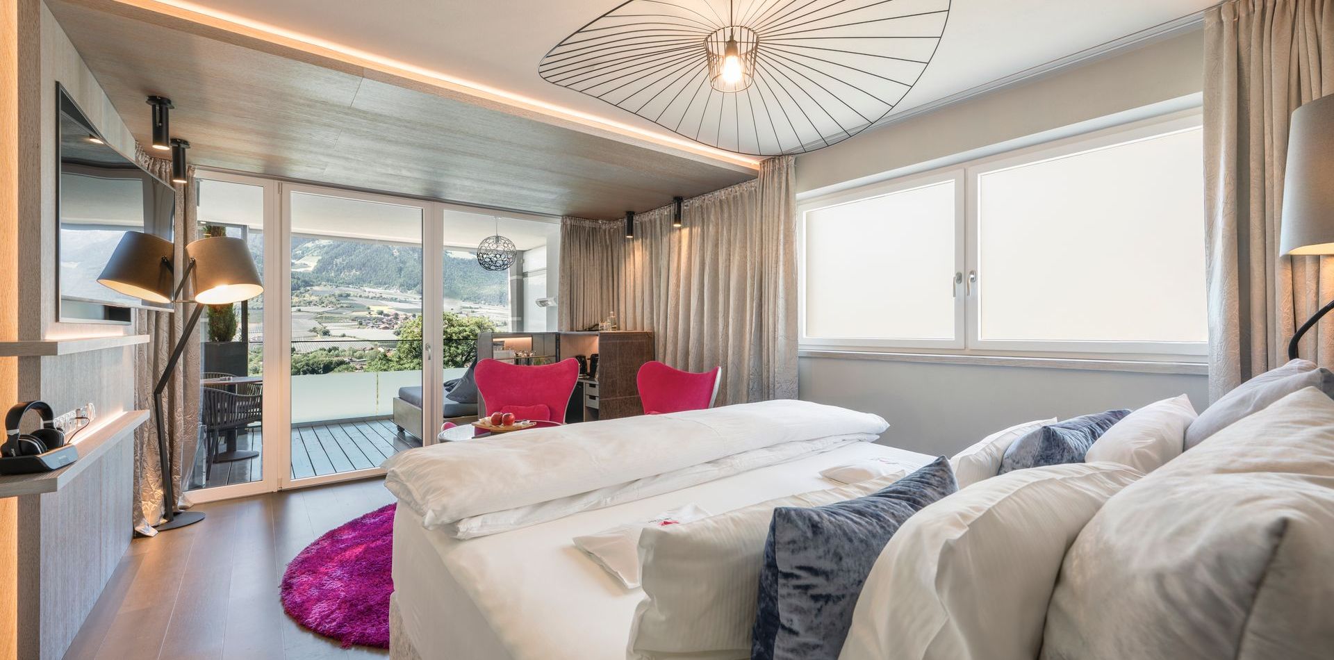 Ein modernes Hotelzimmer mit Doppelbett, pinken Akzenten, Balkon und Blick in die Berge.