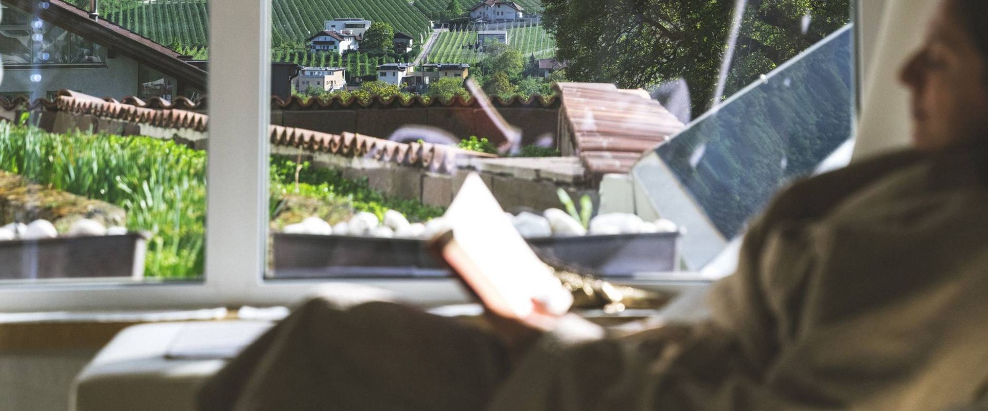 A person relaxes by the window with a book, looking out over vineyard terraces and mountains.