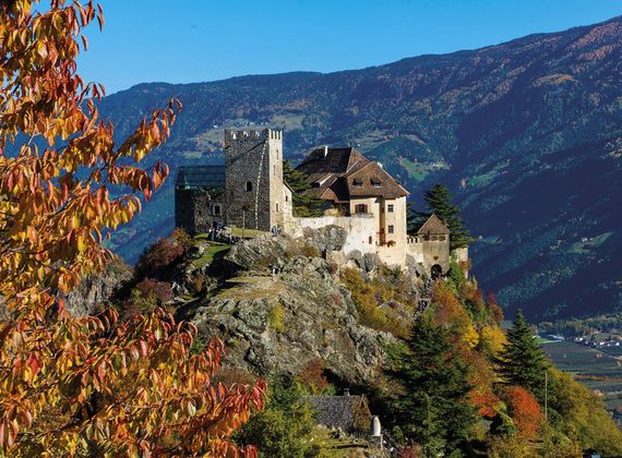 Eine Burg thront auf einem Felsen vor herbstlichen Bäumen und den Bergen im Hintergrund.