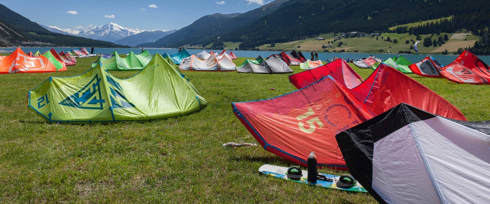 Auf einer Wiese am Reschensee stehen zahlreiche Kitesurf-Zelte vor Bergen und See.