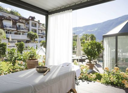 Un lettino per trattamenti si trova su una terrazza luminosa con giardino, hotel e montagne sullo sfondo.
