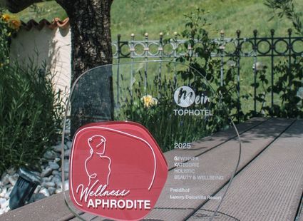 Eine rote Wellness-Trophäe mit der Aufschrift „Wellness Aphrodite“ steht auf einem Holztisch vor grünem Hintergrund.