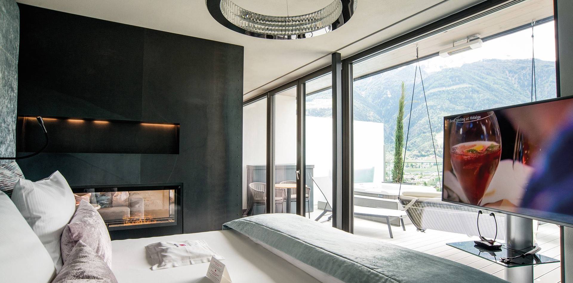 A modern penthouse suite with a large bed, floor-to-ceiling windows, TV and terrace facing the mountains.