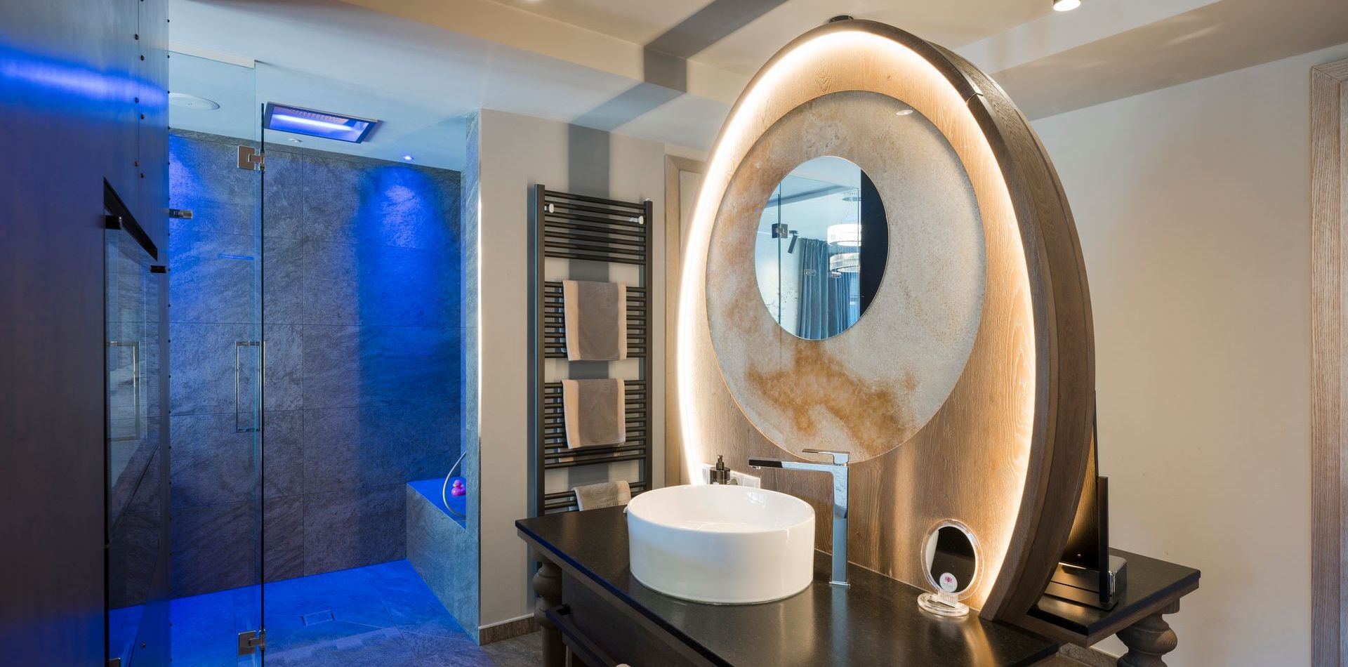 Ein modernes Bad mit Glasdusche, rundem Spiegel und Waschbecken in der Penthouse Suite.