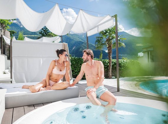 Ein Paar sitzt entspannt an einem kleinen Pool unter einem hellen Sonnensegel mit Bergblick.