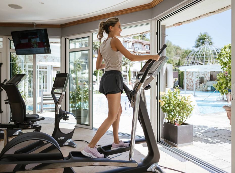 Eine Frau trainiert auf einem Laufband im Fitnessraum mit Blick auf den Poolbereich.