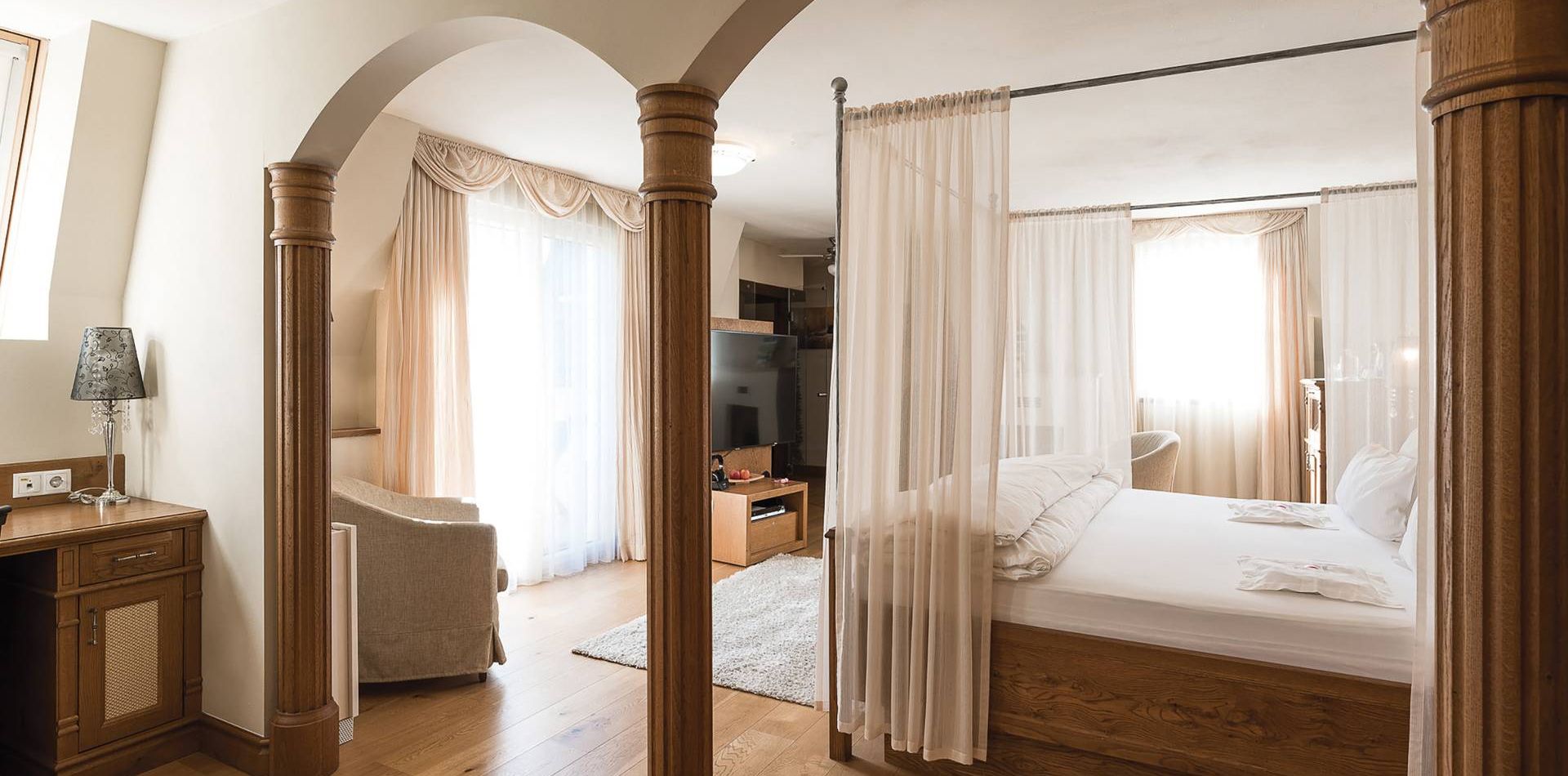 Una spaziosa camera suite con letto a baldacchino, archi in legno e zona salotto in tonalità neutre calde.