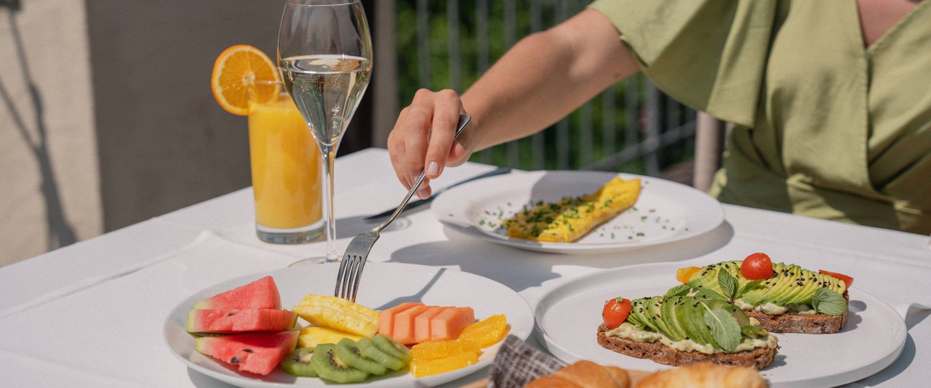 Eine Person greift am gedeckten Tisch zu einem Teller mit Obst, Croissants, Avocado-Toast und einem Getränk.