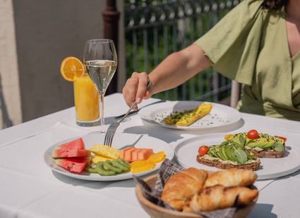 Eine Person greift am gedeckten Tisch zu einem Teller mit Obst, Croissants, Avocado-Toast und einem Getränk.
