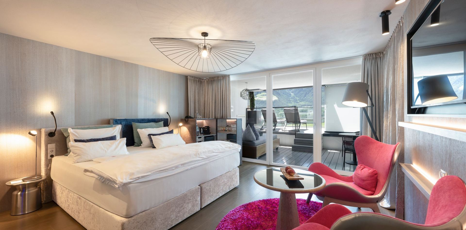 Ein modernes Hotelzimmer mit Doppelbett, rosa Sesseln, runder Tisch und Zugang zur Terrasse.