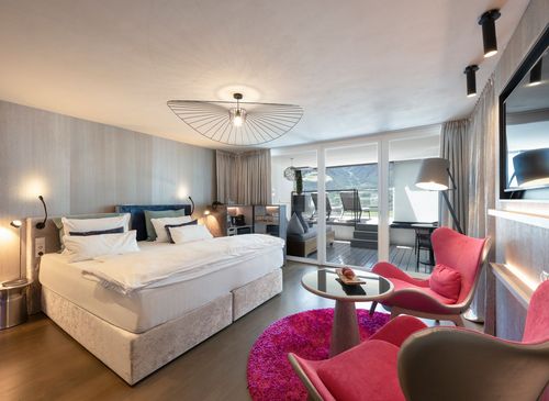 Ein modernes Hotelzimmer mit Doppelbett, rosa Sesseln, runder Tisch und Zugang zur Terrasse.