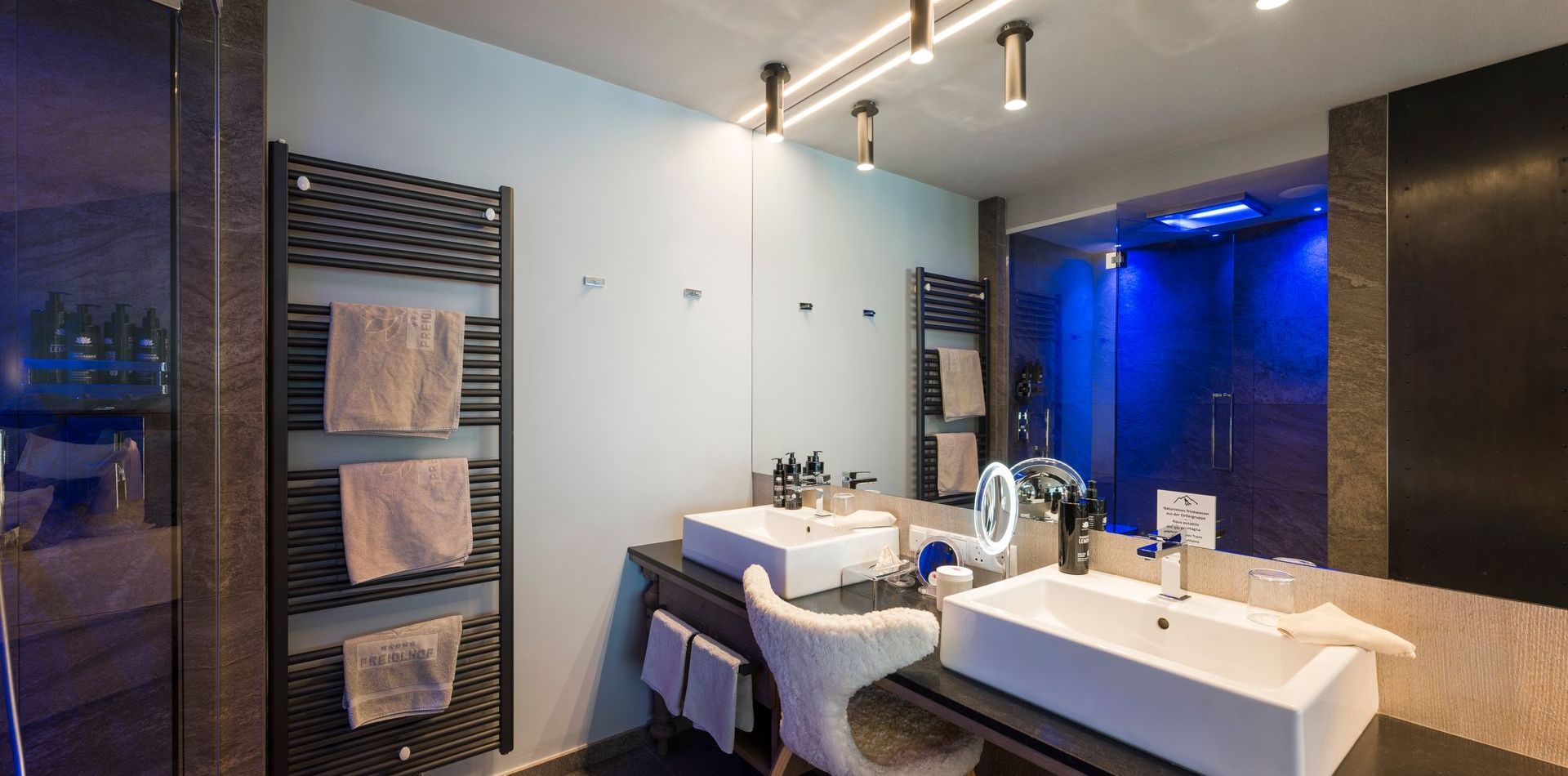 Modernes Badezimmer mit Doppelwaschbecken, beleuchtetem Spiegel, Handtuchheizkörper und dunklen Holzoberflächen im Comfort Room Romantic Fire.