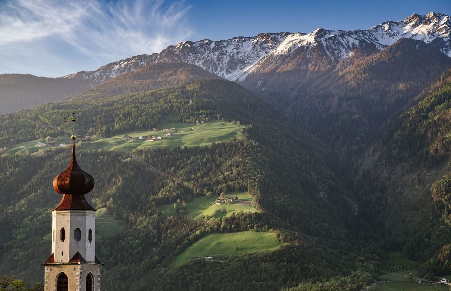 Ein Kirchturm steht vor einer grünen Berglandschaft mit Wiesen und schneebedeckten Gipfeln.