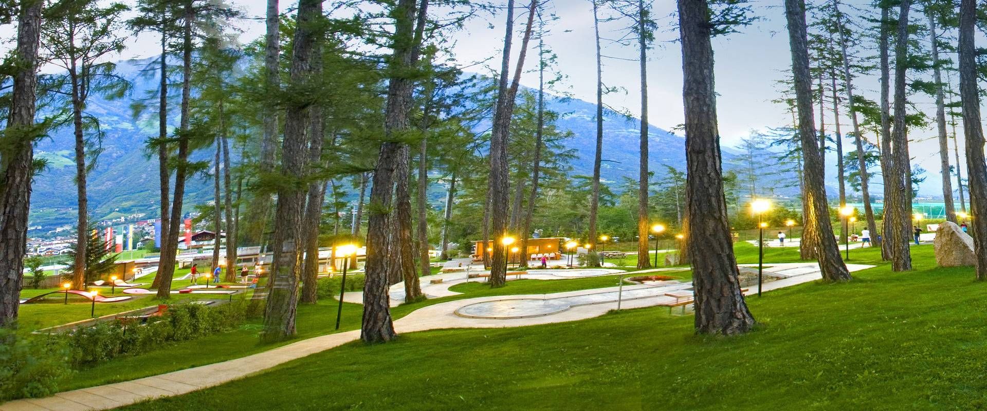 Zwischen hohen Bäumen liegt eine Minigolfanlage auf einer gepflegten Wiese mit Bergkulisse im Hintergrund.