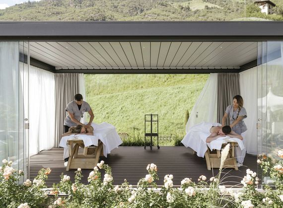 Zwei Gäste erhalten im offenen Spa-Bereich gleichzeitig eine Massage auf zwei Liegen mit Blick ins Grüne.