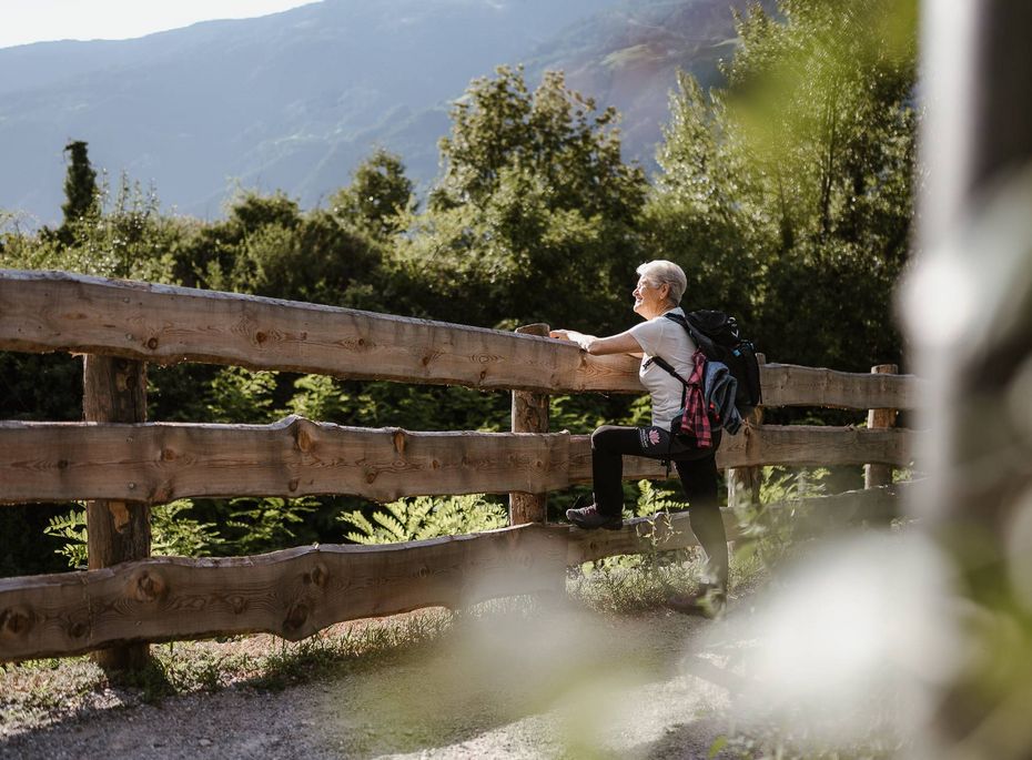Wandern am Preidlhof in Südtirol Eine ältere Frau sitzt mit Rucksack an einem Holzzaun und blickt auf die Berge.