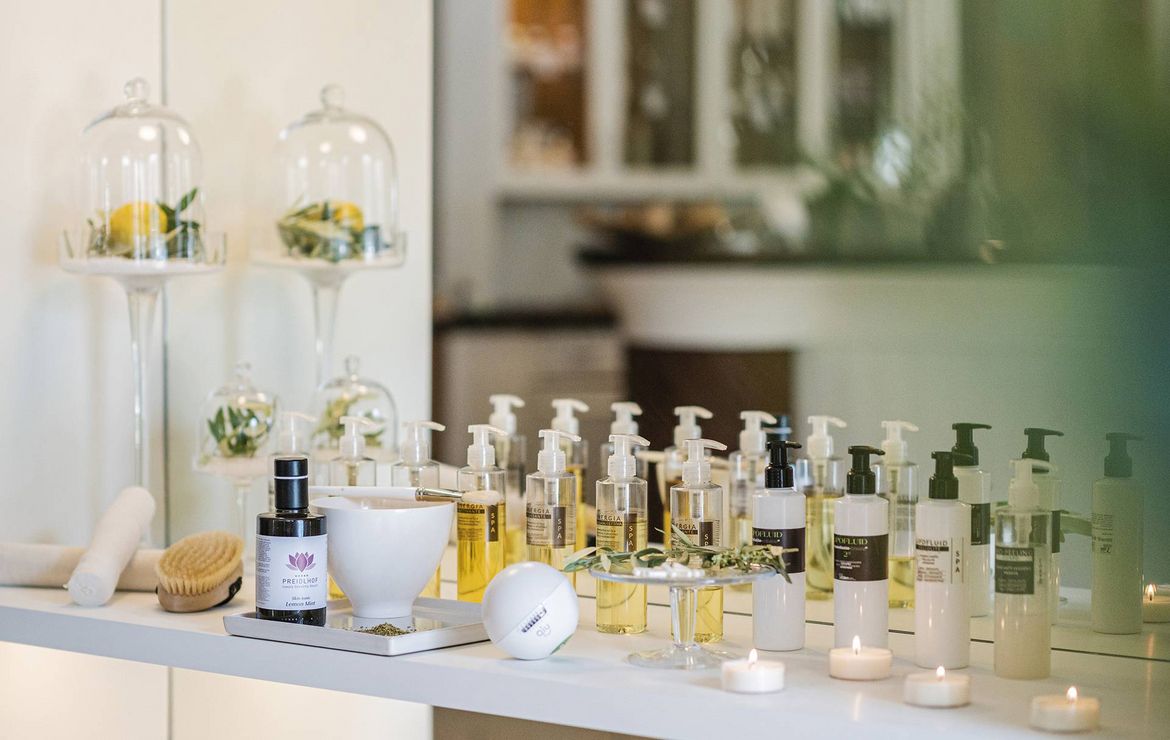 A row of spa treatment products and dispensers is arranged on a white beauty counter with candles and glass cloches.