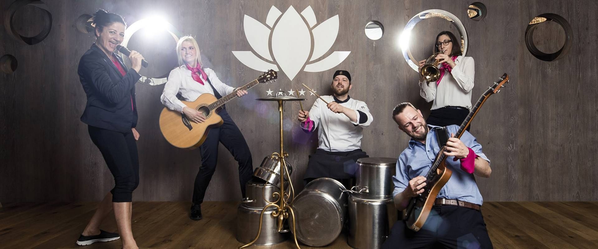 Vier Mitarbeitende des Preidlhof posieren mit Instrumenten in einem Raum mit Holzfußboden und dekorativer Wand.