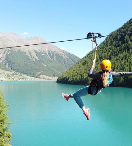 Eine Person gleitet an einer Seilrutsche über einen türkisfarbenen Bergsee zwischen bewaldeten Hängen.