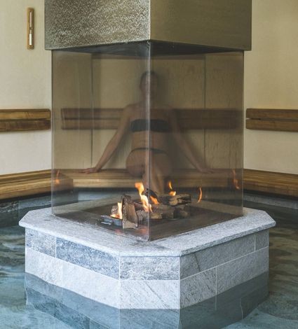 A person sits behind a glass-enclosed fireplace inside an indoor sauna.
