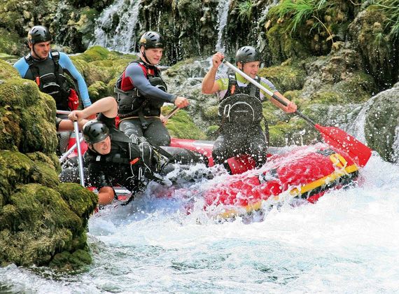 Vier Personen navigieren mit einem Schlauchboot durch wilde Stromschnellen zwischen Felsen und Wasser.