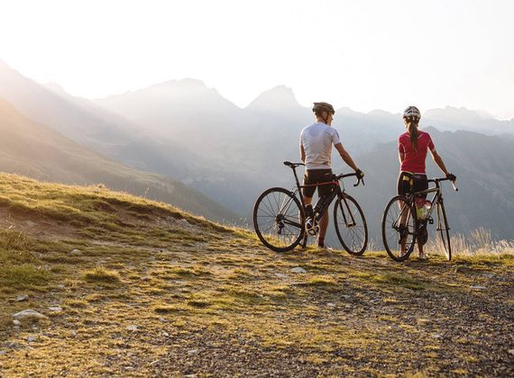 Zwei Rennradfahrer stehen auf einer Wiese und blicken in die Berglandschaft im Abendlicht.