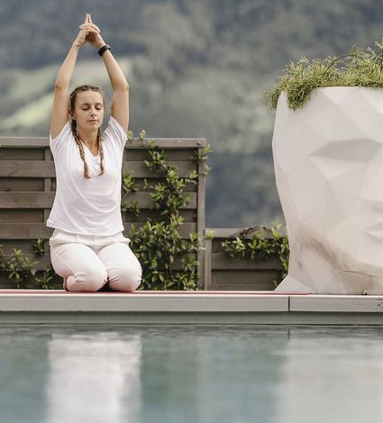 A woman kneels on a mat by the pool with her arms raised overhead in a yoga pose.