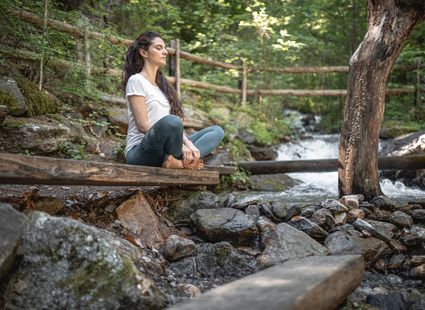 Eine Frau sitzt auf einer Holzplattform am Bach und meditiert im Wald.