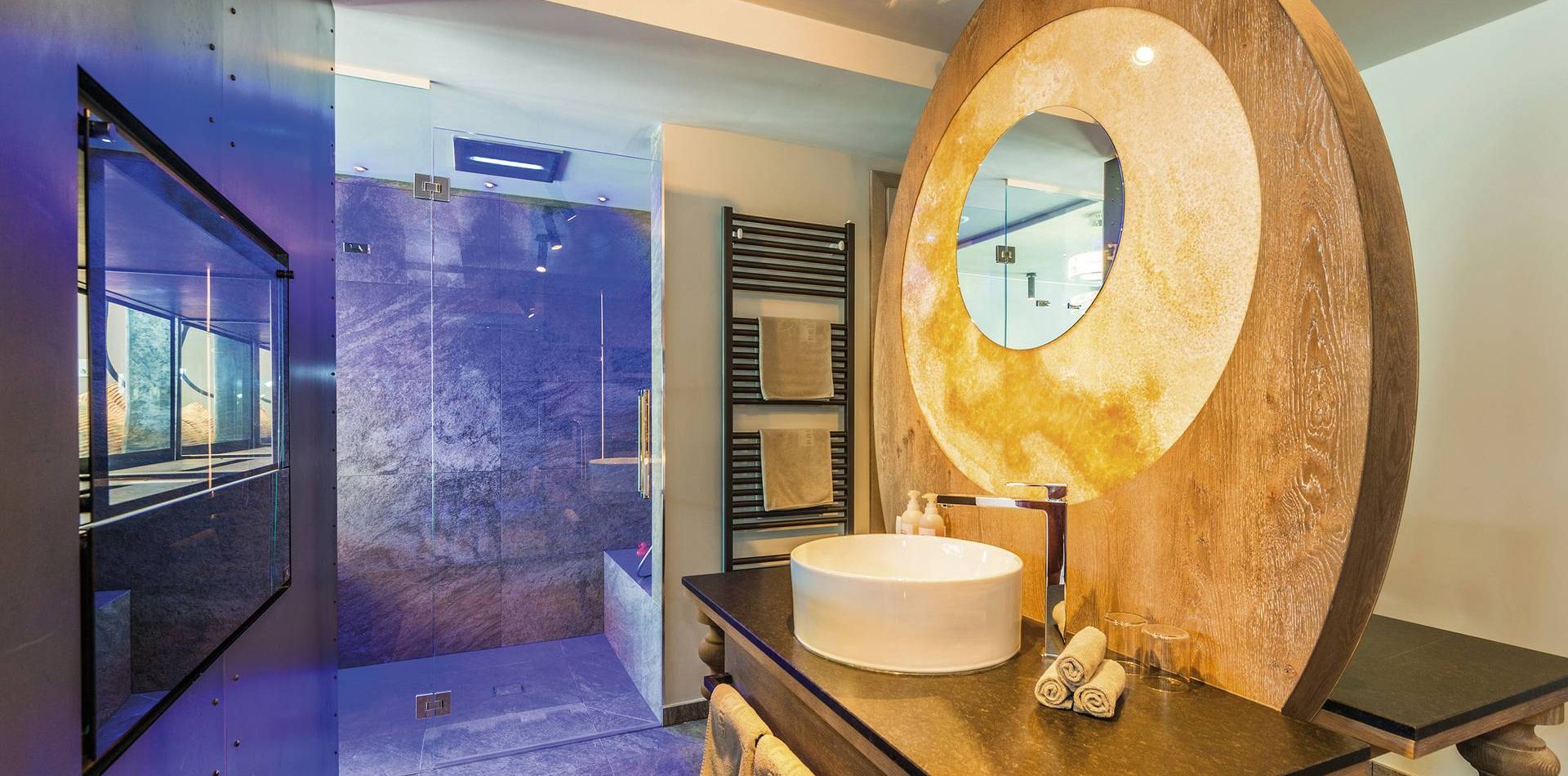 A modern bathroom in the penthouse suite with a circular wood-framed mirror, vessel sink, and walk-in shower.