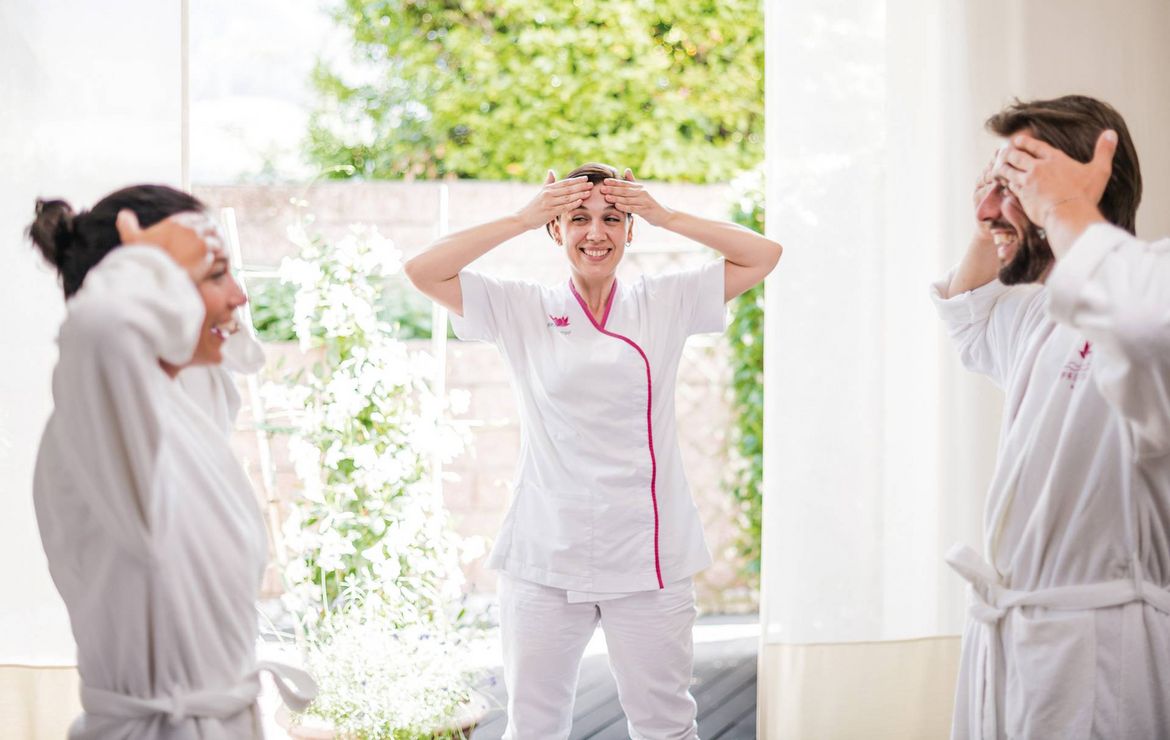 Drei Personen im Bademantel führen eine Wellness-Übung mit Handbewegungen in einem hellen Raum aus.