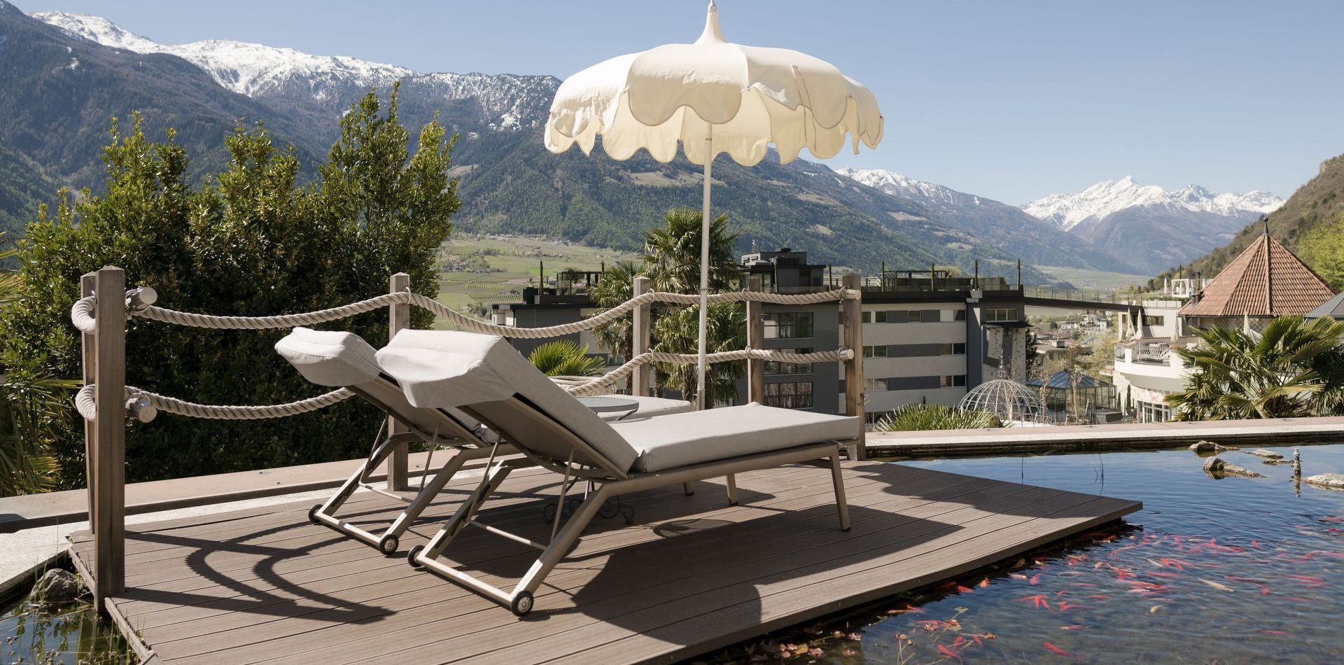 Eine Holzterrasse mit zwei Liegen, Sonnenschirm und Naturpool vor Bergpanorama bei der Luxury Suite Lantana.