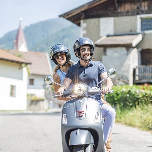 Ein Paar fährt mit einem Roller durch ein Dorf in Südtirol, im Hintergrund liegen Berge und Häuser am Straßenrand.