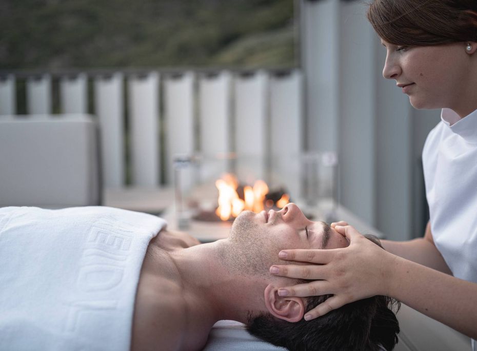Eine Therapeutin behandelt das Gesicht eines liegenden Mannes in einem hellen Spa-Raum.