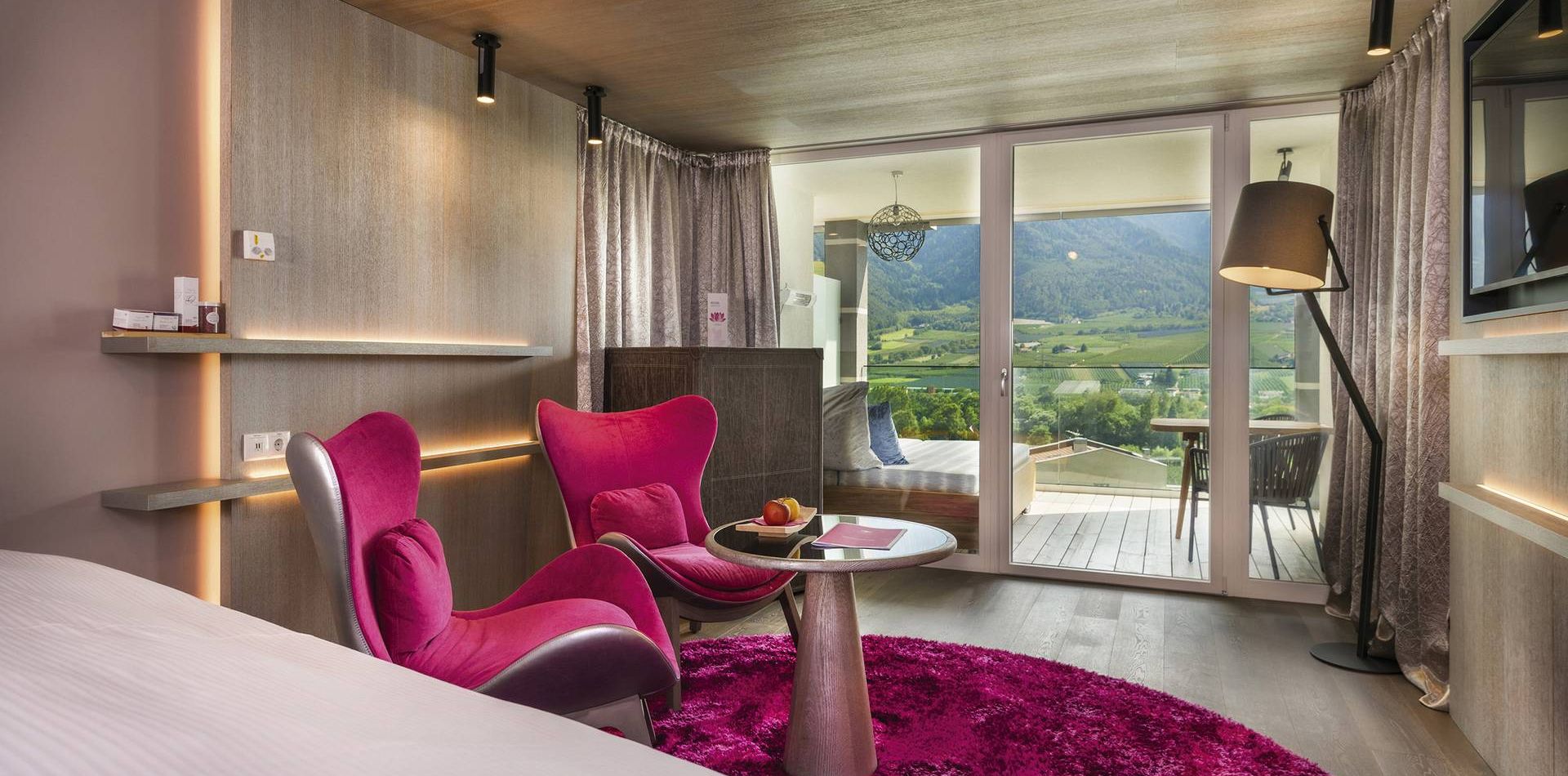 Una luxury suite con sedute fucsia, tavolino rotondo, bordo del letto e porte sul balcone con vista sulle montagne.
