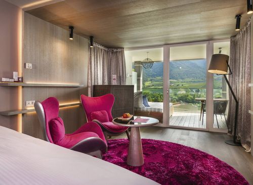 A luxury suite with magenta chairs, a round table, a bed edge and balcony doors overlooking the mountains.