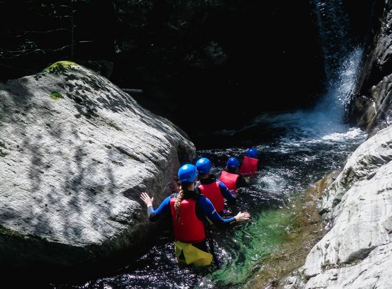 Drei Personen mit Helmen und Schwimmwesten waten durch eine enge Schlucht mit Wasserlauf.