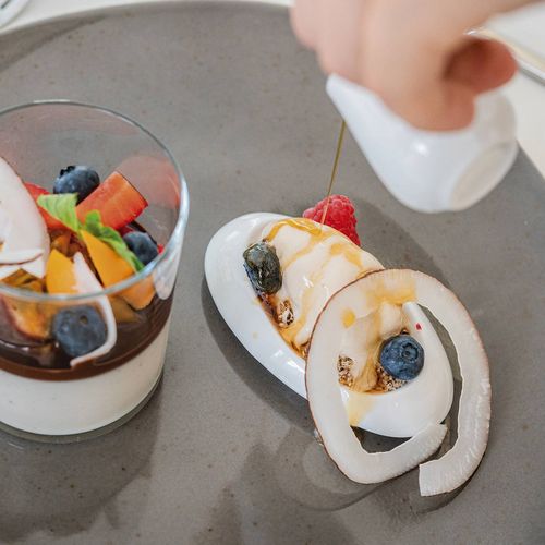 Ein Dessert mit Früchten und Beeren wird auf einem grauen Teller angerichtet.