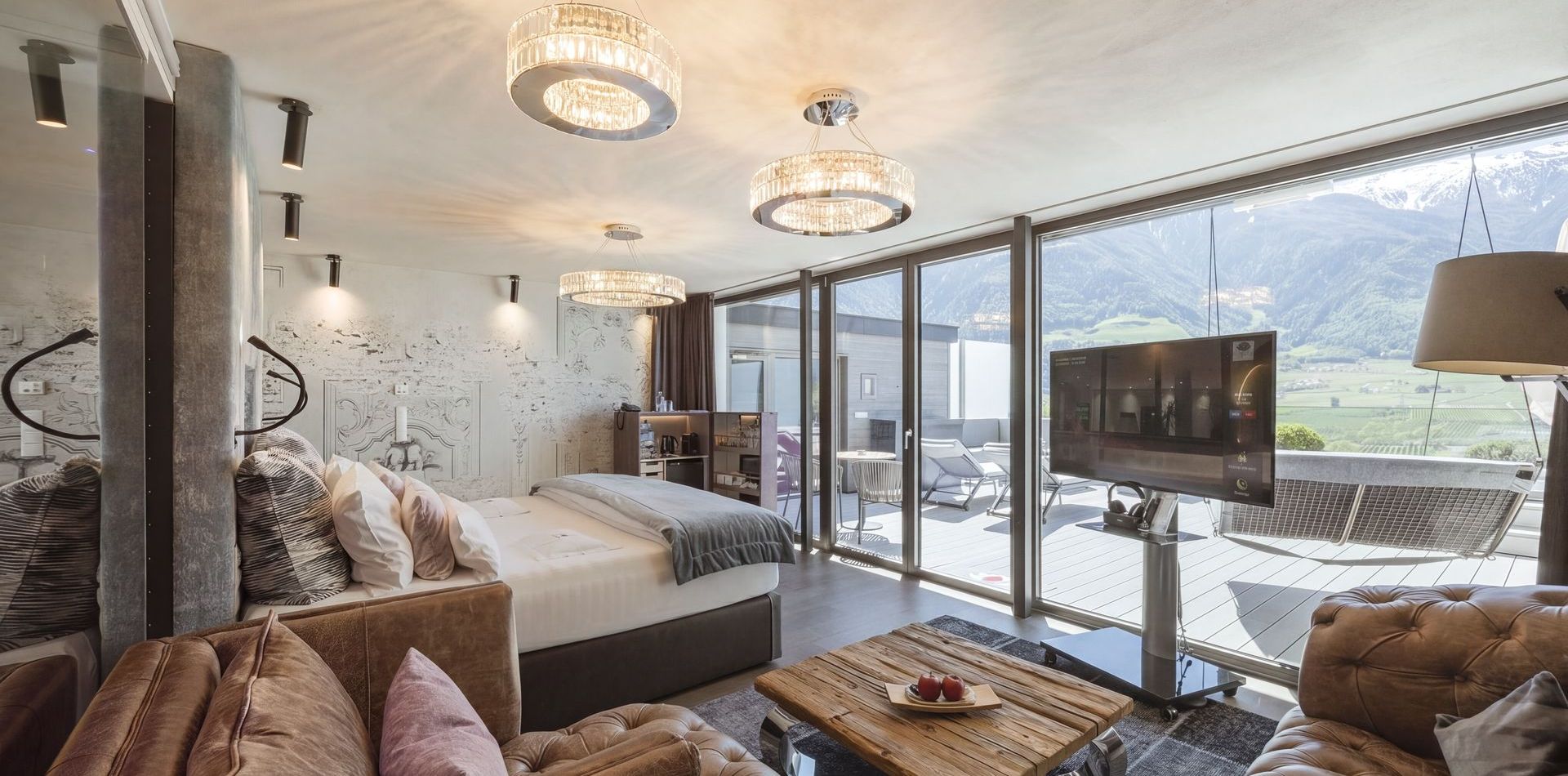 Ein modernes Schlafzimmer der Penthouse Suite mit Sofa, Holztisch und großer Glasfront zum Balkon mit Bergblick.