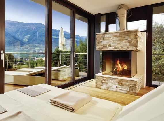 Ein heller Ruheraum mit zwei Liegen, großem Kamin und Blick auf die Berge durch bodentiefe Fenster.